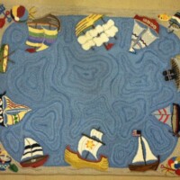 A colorful nautical-themed rug with various maritime elements such as boats, a lighthouse, sea creatures, and nautical flags, set against a blue wavy background.