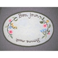 Oval-shaped decorative mat with the phrase "bon jour" surrounded by floral embroidery, viewed upside down.