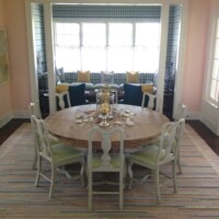 A spacious dining room with a large oval table, white chairs, and two blue armchairs set on a multicolored striped rug, with natural light filtering through checkered window curtains.