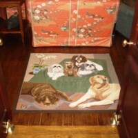 A doormat with dog illustrations in front of a wooden door, flanked by furniture with red floral patterns.