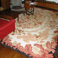 A living room with a floral patterned area rug, a red armchair, and a small table with items on it, featuring hardwood floors and a fireplace.