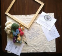 Rug making materials and pattern set out on a table, including a stretcher, monks cloth, puncher, and colored yarn.