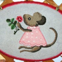 Handcrafted textile featuring an anthropomorphic mouse in a pink dress holding a flower, displayed in an oval frame.