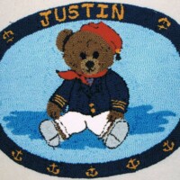 An embroidered mat featuring an illustration of a teddy bear wearing a sailor outfit with the name "justin" above it.