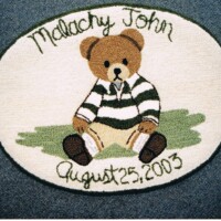 A personalized rug featuring an illustration of a teddy bear wearing a striped shirt with the name "malachy john" and the date "august 25, 2003" embroidered on it.